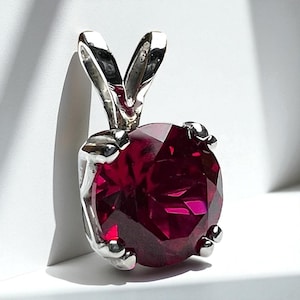 May include: A silver pendant with a large, faceted, deep red gemstone. The pendant has a bail with a loop design. The gemstone is round and has multiple facets that reflect light. The pendant is set against a white background.