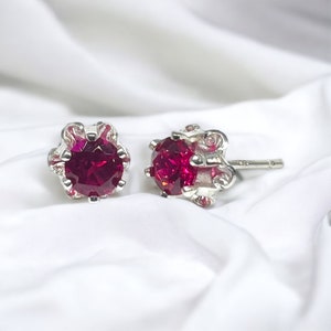 May include: A pair of silver stud earrings with a red gemstone in the center of each earring. The earrings have a detailed design around the gemstone.