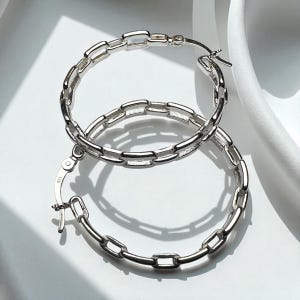 May include: A pair of silver hoop earrings with a chain link design. The earrings are made of a shiny metal and have a clasp closure. The earrings are displayed on a white surface with soft shadows.