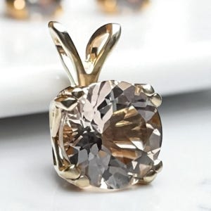 May include: A gold pendant with a large, faceted, light brown gemstone. The gemstone is square-cut and set in a gold setting with a decorative bail. The pendant is displayed on a white surface.