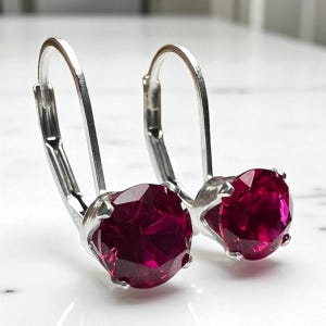May include: A pair of silver leverback earrings, each featuring a large, faceted, round, deep red gemstone. The earrings are displayed on a white surface, showcasing the vibrant color and sparkle of the stones.