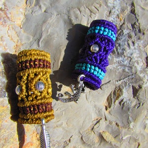 Puede incluir: Dos pulseras de macramé hechas a mano con abalorios de metal plateado. Una pulsera es morada con detalles turquesa y un abalorio de lagarto. La otra pulsera es marrón y dorada con un abalorio de pluma.