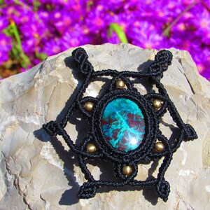 May include: A black macrame pendant with a green and red gemstone in the center. The pendant is surrounded by gold beads and is resting on a gray rock.