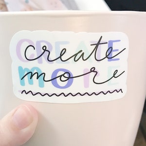 May include: A pink ceramic mug with a white sticker that says "create more" in black script. The sticker has a colorful, abstract design with purple, blue, and green accents.