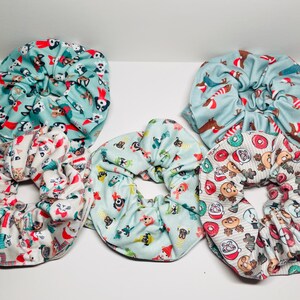 May include: Five colorful scrunchies with holiday-themed patterns. The scrunchies are made of soft fabric and feature various designs, including dogs, donuts, and Christmas trees.