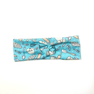 May include: A light blue headband with a pattern of baking tools and white polka dots. The headband is knotted in the center.