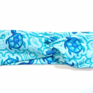 Op de afbeelding: Een blauw en turquoise hoofdband met een gedraaid ontwerp heeft een herhalend patroon van blauwe zeeschildpadden.