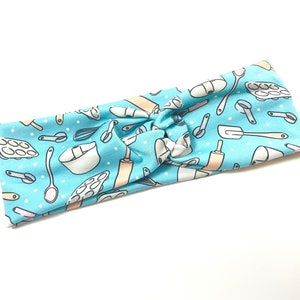 May include: A light blue headband with a pattern of baking tools and white polka dots. The tools include a rolling pin, whisk, measuring cups, a spatula, and a mixing bowl.
