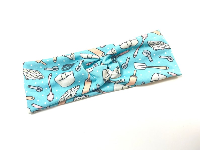 May include: A light blue headband with a pattern of baking tools and white polka dots. The tools include a rolling pin, whisk, measuring cups, a spatula, and a mixing bowl.