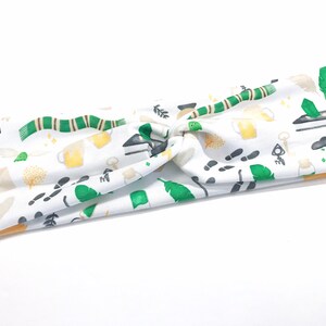 May include: White knotted headband with a green, gold, and black print of various Harry Potter themed icons, including a scarf, a feather, a key, and a cup.