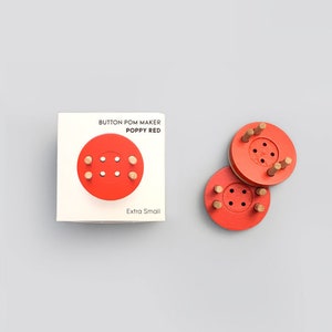 May include: A set of poppy red extra small button pom makers. The makers are circular with wooden pegs and a central hole. The packaging is a white box with the text "BUTTON POM MAKER POPPY RED Extra Small".