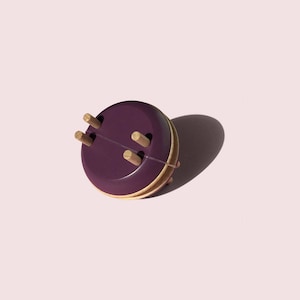 May include: A round, purple coaster with four wooden pegs. The coaster has a gold-colored rim and is set against a light pink background. The coaster is designed to protect surfaces from heat and moisture.