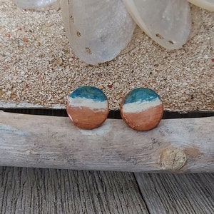 May include: A pair of round copper earrings with a blue, white, and copper layered design. The earrings are displayed on a piece of driftwood.