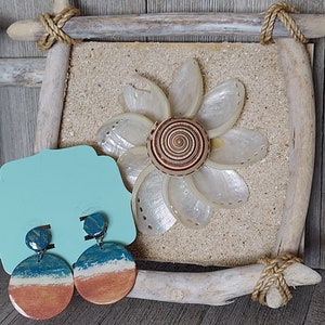 Puede incluir: Un par de pendientes redondos de madera con un diseño de pintura azul, blanco y naranja. Los pendientes se muestran en una tarjeta azul claro con un fondo de flor de concha.