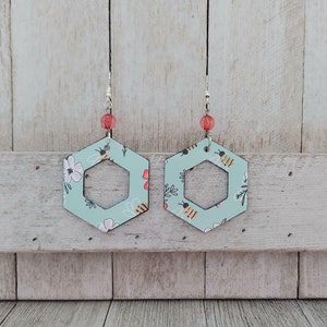 May include: A pair of hexagonal earrings with a light blue background and a pattern of bees and flowers. The earrings have a silver hook and a red bead at the top.