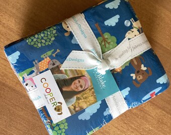 May include: A stack of folded fabric squares featuring a blue background with a repeating pattern of cartoon dogs, trees, and clouds. The stack is tied with a white ribbon printed with the text Riley Blake Designs and includes a Cooper label.
