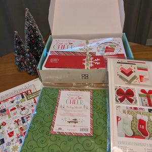 May include: A Christmas-themed crafting kit with a green fabric featuring a swirl pattern, a red and white striped label, and the text "Cup of Cheer". The kit includes a box, instructions, and decorative elements.