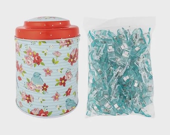 May include: A cylindrical metal tin with a red lid and a light blue body featuring a floral and bird pattern, accompanied by a clear plastic bag filled with numerous translucent light blue plastic sewing clips with metal springs.