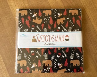 May include: A square stack of fabric squares wrapped in clear plastic, featuring a black background with a repeating pattern of brown bears, red pine trees, campfires, axes, saws, and lanterns. A white paper band reads WOODSMAN by Lori Whitlock.