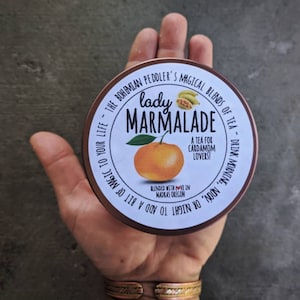 Puede incluir: Una lata redonda de metal marr&oacute;n con una etiqueta blanca que dice "lady marmalade" en fuente negra. La etiqueta tambi&eacute;n incluye una peque&ntilde;a naranja, algunas hojas verdes y el texto "A tea for cardamom lovers!" en fuente negra. El texto "BLENDED WITH LOVE IN MADRAS OREGON" tambi&eacute;n est&aacute; en la etiqueta en fuente negra. La lata de metal est&aacute; etiquetada "THE BOHEMIAN PEDDLER'S MAGICAL BLENDS OF TEA - DRINK MORNING, NOON, OR NIGHT TO ADD A BIT OF MAGIC TO YOUR LIFE."