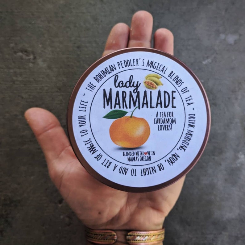 Puede incluir: Una lata redonda de metal marr&oacute;n con una etiqueta blanca que dice "lady marmalade" en fuente negra. La etiqueta tambi&eacute;n incluye una peque&ntilde;a naranja, algunas hojas verdes y el texto "A tea for cardamom lovers!" en fuente negra. El texto "BLENDED WITH LOVE IN MADRAS OREGON" tambi&eacute;n est&aacute; en la etiqueta en fuente negra. La lata de metal est&aacute; etiquetada "THE BOHEMIAN PEDDLER'S MAGICAL BLENDS OF TEA - DRINK MORNING, NOON, OR NIGHT TO ADD A BIT OF MAGIC TO YOUR LIFE."