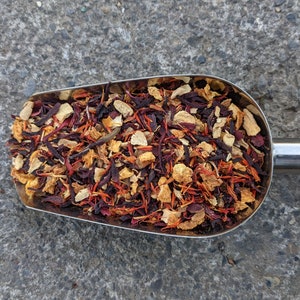 May include: A stainless steel scoop filled with a colorful blend of dried tea leaves and herbs. The mix includes deep red hibiscus, orange pieces, and light brown ginger, creating a visually appealing and aromatic product.