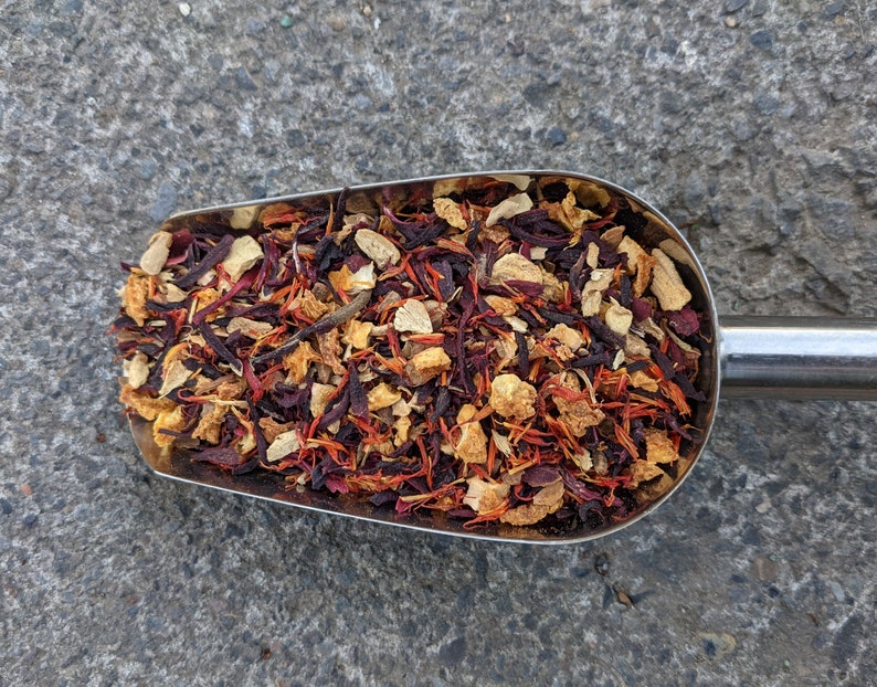 May include: A stainless steel scoop filled with a colorful blend of dried tea leaves and herbs. The mix includes deep red hibiscus, orange pieces, and light brown ginger, creating a visually appealing and aromatic product.