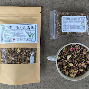 May include: A brown paper bag with a white label that reads "Holy Basil Transition Tea" and lists the ingredients: tulsi leaf, chamomile, rosehips, lemongrass, orange peel, lemon peel, jasmine, and rose petals. The label also includes instructions for steeping the tea and a note that it is blended with love in Madras, Oregon. A small clear plastic bag filled with the tea blend is visible in the background.