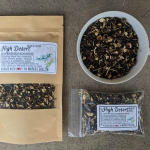 May include: A brown paper bag with a label that reads "High Desert Breakfast Blend" and contains a blend of black tea, juniper berries, ginger, allspice, rosemary, and cardamom seeds. The bag is sitting on a gray concrete surface.