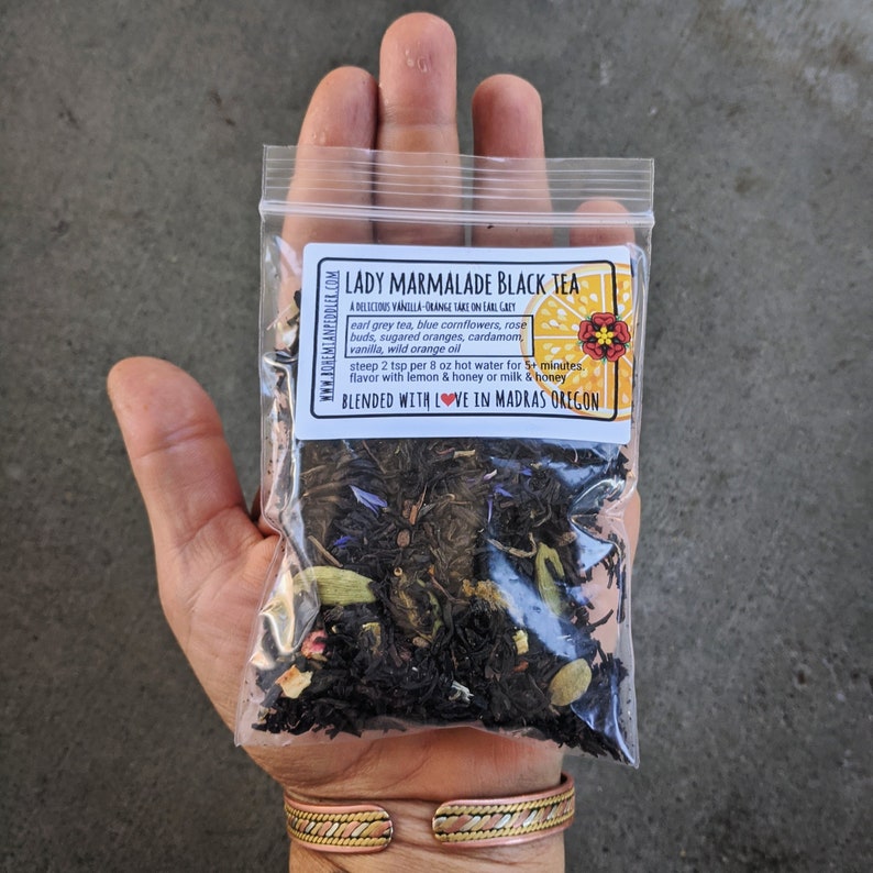 Puede incluir: Una peque&ntilde;a bolsa de pl&aacute;stico transparente llena de t&eacute; negro suelto llamado "Lady Marmalade Black Tea". La mezcla de t&eacute; incluye t&eacute; Earl Grey, aciano azul, capullos de rosa, naranjas azucaradas, cardamomo, vainilla y aceite de naranja silvestre. La bolsa tiene una etiqueta con el texto "Una deliciosa toma de vainilla y naranja en Earl Grey" y "Mezclado con amor en Madras Oreg&oacute;n".