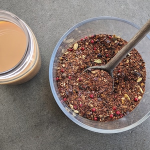 Op de afbeelding: Een glazen kom gevuld met losse theeblend, waaronder rode rooibos, gedroogde fruitstukjes en specerijen. Een houten lepel is gedeeltelijk ondergedompeld in de theeblend.