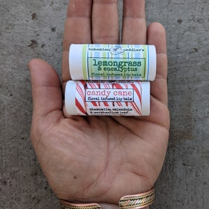 May include: Two small tubes of lip balm, one with a red and white candy cane design and the text "candy cane" and the other with a green and white design and the text "lemongrass & eucalyptus". Both tubes have the text "bohemian peddler's" and "floral infused lip balm".
