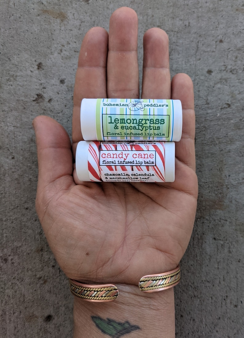 May include: Two small tubes of lip balm, one with a red and white candy cane design and the text "candy cane" and the other with a green and white design and the text "lemongrass & eucalyptus". Both tubes have the text "bohemian peddler's" and "floral infused lip balm".