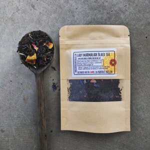 Puede incluir: Una bolsa de papel marr&oacute;n con una ventana transparente que muestra t&eacute; negro de hoja suelta. La bolsa tiene una etiqueta que dice "Lady Marmalade Black Tea" y enumera los ingredientes: t&eacute; earl grey, aciano azul, capullos de rosa, naranjas azucaradas, cardamomo, vainilla, aceite de naranja silvestre. La etiqueta tambi&eacute;n incluye instrucciones para preparar 2 cucharaditas por 225 ml de agua caliente durante 15 minutos y aromatizar con lim&oacute;n y miel o leche y miel. La etiqueta termina con "Blended with Love in Madras Oregon."
