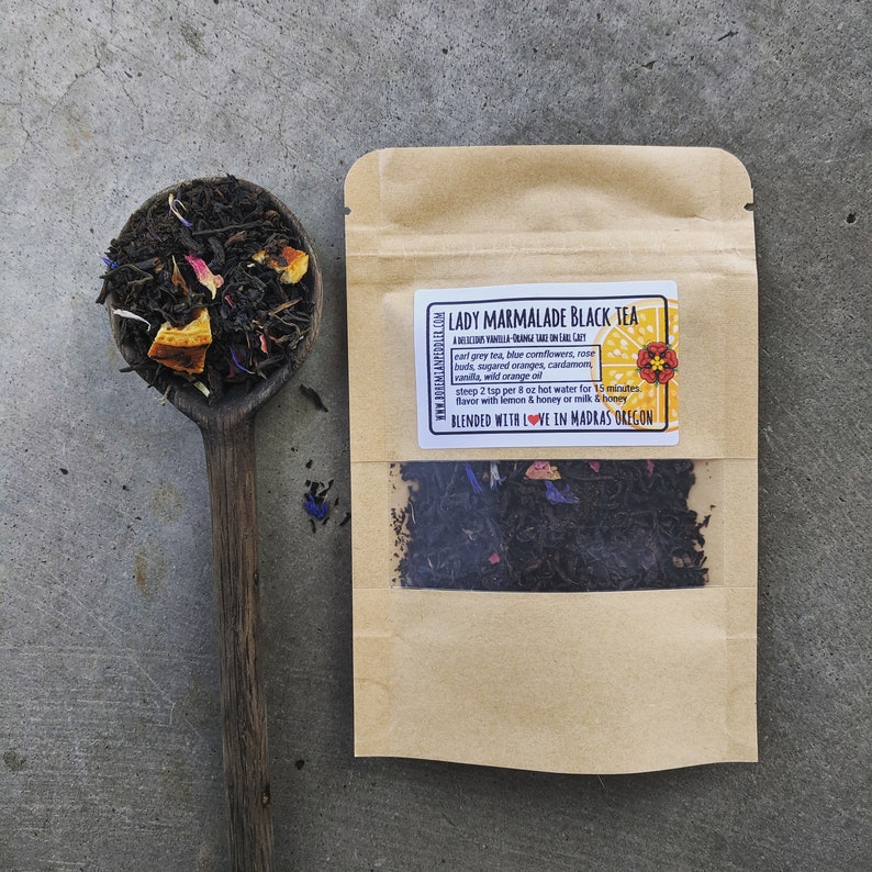 Puede incluir: Una bolsa de papel marr&oacute;n con una ventana transparente que muestra t&eacute; negro de hoja suelta. La bolsa tiene una etiqueta que dice "Lady Marmalade Black Tea" y enumera los ingredientes: t&eacute; earl grey, aciano azul, capullos de rosa, naranjas azucaradas, cardamomo, vainilla, aceite de naranja silvestre. La etiqueta tambi&eacute;n incluye instrucciones para preparar 2 cucharaditas por 225 ml de agua caliente durante 15 minutos y aromatizar con lim&oacute;n y miel o leche y miel. La etiqueta termina con "Blended with Love in Madras Oregon."