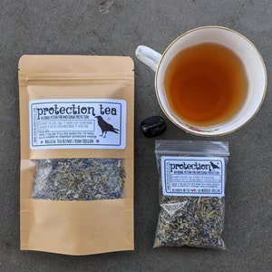 May include: A brown paper bag with a clear window showing a blend of dried herbs. The bag has a label that reads "protection tea" and includes the text "An herbal potion for emotional protection." The bag is sitting on a gray surface next to a small clear bag with a similar label and a cup of tea.