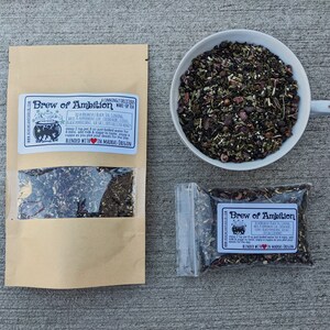 May include: Two brown paper bags with clear plastic windows showing loose leaf tea inside. The bags are labeled "Brew of Ambition" with a black cauldron graphic and text describing the tea blend. The text includes "Cunningly Delicious Black Tea" and "Blended with Love in Madras Oregon". A white ceramic cup with a handle is filled with the loose leaf tea.