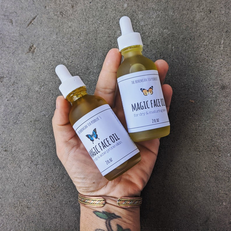 May include: Two amber glass bottles with dropper tops, labeled "Magic Face Oil" for different skin types. The bottles are held in a hand with a gold bracelet.