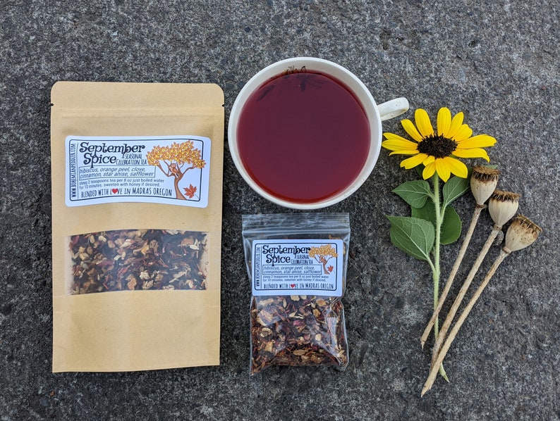 May include: An overhead shot of a cup of red tea, two pouches of tea, a sunflower, and dried seed pods. The tea pouches are labeled "September Spice" with a list of ingredients. The tea cup is white and the tea is a deep red color.