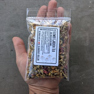 May include: A small clear plastic bag filled with a blend of dried flowers and herbs for making tea. The bag has a white label with the text "Flower Garden Tea" and instructions for brewing the tea. The label also includes the website address "www.bohemianpeddler.com" and the text "Blended with love in Madras Oregon".