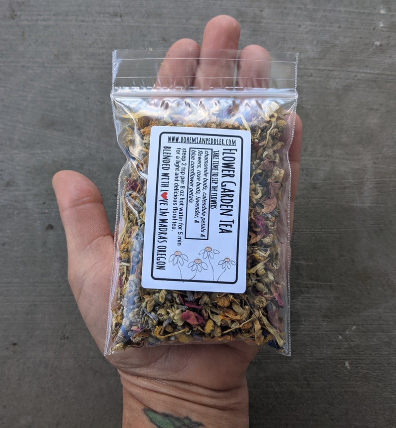 May include: A small clear plastic bag filled with a blend of dried flowers and herbs for making tea. The bag has a white label with the text "Flower Garden Tea" and instructions for brewing the tea. The label also includes the website address "www.bohemianpeddler.com" and the text "Blended with love in Madras Oregon".