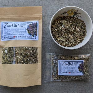May include: Two brown paper bags with a white label that reads "Zen ERGY A CRUEL ENERGY BOOST FOR YOUR BODY & MIND" and "BLENDED WITH LOVE IN MADRAS OREGON". The bags contain dried herbs. A white teacup with a floral pattern is filled with the dried herbs.