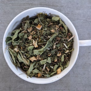 May include: A white ceramic bowl filled with a blend of dried herbs and fruit pieces. The mixture includes green leaves, small pieces of dried fruit, and other botanicals.