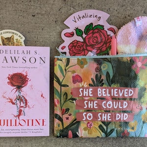 May include: A pink book titled "Guillotine" by Delilah S. Dawson, with a cat bookmark. A floral cosmetic bag with the text "She Believed She Could So She Did" and a pink floral design. Includes a pink face mask.