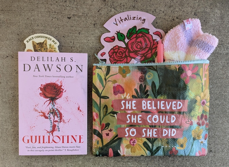 May include: A pink book titled "Guillotine" by Delilah S. Dawson, with a cat bookmark. A floral cosmetic bag with the text "She Believed She Could So She Did" and a pink floral design. Includes a pink face mask.