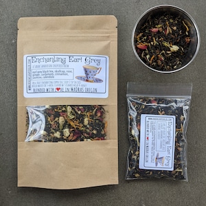 May include: Two bags of Enchanting Earl Grey tea blend, a Sarah Sanderson inspired brew. The tea blend includes earl grey black tea, skullcap, rose, ginger, cardamom, cinnamon, jasmine, and calendula. The bags are on a gray surface with a small metal bowl of loose leaf tea.