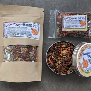 May include: A brown paper bag with a clear window showing a mix of dried spices and fruit peels. The bag has a label that reads "Autumn Magic Mulling Spice" and includes a list of ingredients. The bag is next to a small clear plastic bag with a similar mix of spices and a tin with a similar label.