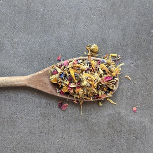 May include: A wooden spoon filled with a mixture of dried flowers, including yellow, pink, and purple petals. The flowers are arranged in a loose, natural way, creating a colorful and fragrant blend.