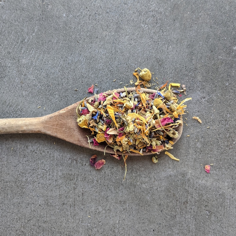 May include: A wooden spoon filled with a mixture of dried flowers, including yellow, pink, and purple petals. The flowers are arranged in a loose, natural way, creating a colorful and fragrant blend.