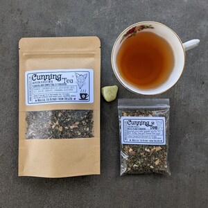 May include: A brown paper bag with a white label that reads "Cunning Tea" and a fox illustration. The label also includes a list of ingredients and instructions for brewing the tea. The bag contains loose leaf tea. A small clear plastic bag with a similar label and loose leaf tea is also pictured. A cup of tea and a green stone are in the background.